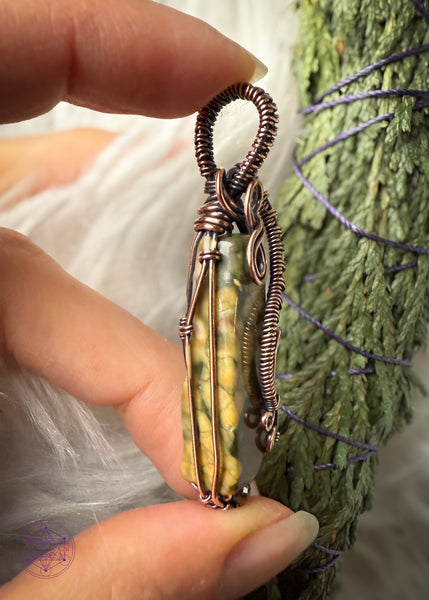 Rainforest Rhyolite Pendant wrapped in copper wire. The copper curls around the face of the stone to show the details of the green and brown in the crystal, and adding curls at the bottom with the wire like fern curls. The base of the pendant I added the Green tourmaline bead to fill in the point I added at the bottom with wire. The bail is simple narrow weave. Held showing the left side and the thickness.