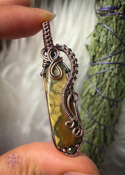 Rainforest Rhyolite Pendant wrapped in copper wire. The copper curls around the face of the stone to show the details of the green and brown in the crystal, and adding curls at the bottom with the wire like fern curls. The base of the pendant I added the Green tourmaline bead to fill in the point I added at the bottom with wire. The bail is simple narrow weave. Held in hand with a white faux fur behind it with a cedar bundle to tie in the forest look.