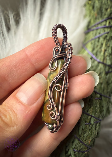 Rainforest Rhyolite Pendant wrapped in copper wire. The copper curls around the face of the stone to show the details of the green and brown in the crystal, and adding curls at the bottom with the wire like fern curls. The base of the pendant I added the Green tourmaline bead to fill in the point I added at the bottom with wire. The bail is simple narrow weave. Showing the right side details.