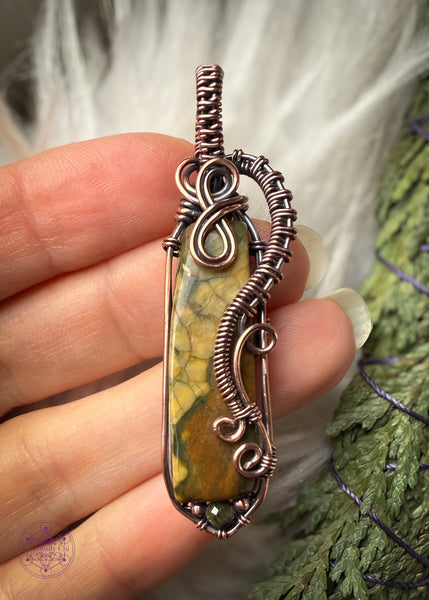 Rainforest Rhyolite Pendant wrapped in copper wire. The copper curls around the face of the stone to show the details of the green and brown in the crystal, and adding curls at the bottom with the wire like fern curls. The base of the pendant I added the Green tourmaline bead to fill in the point I added at the bottom with wire. The bail is simple narrow weave. Held in hand for size reference (four finger widths wide) with a white faux fur behind it with a cedar bundle to tie in the forest look.