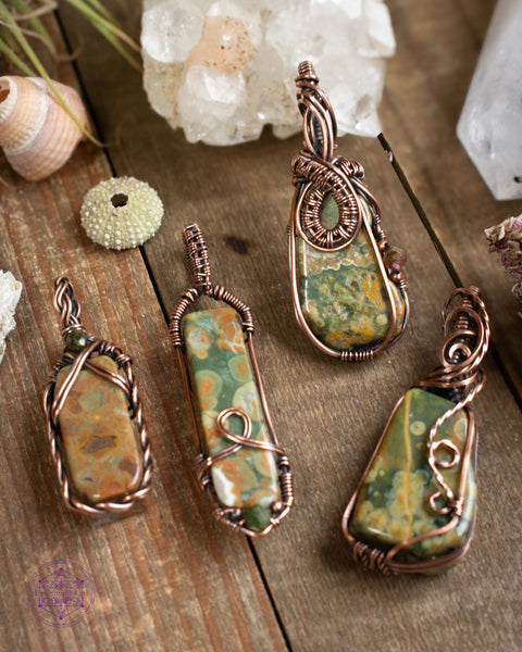 copper wire wrapped rainforest rhyolite pendant collection. four pendants sitting on a wooden table with crystals and shells around it.