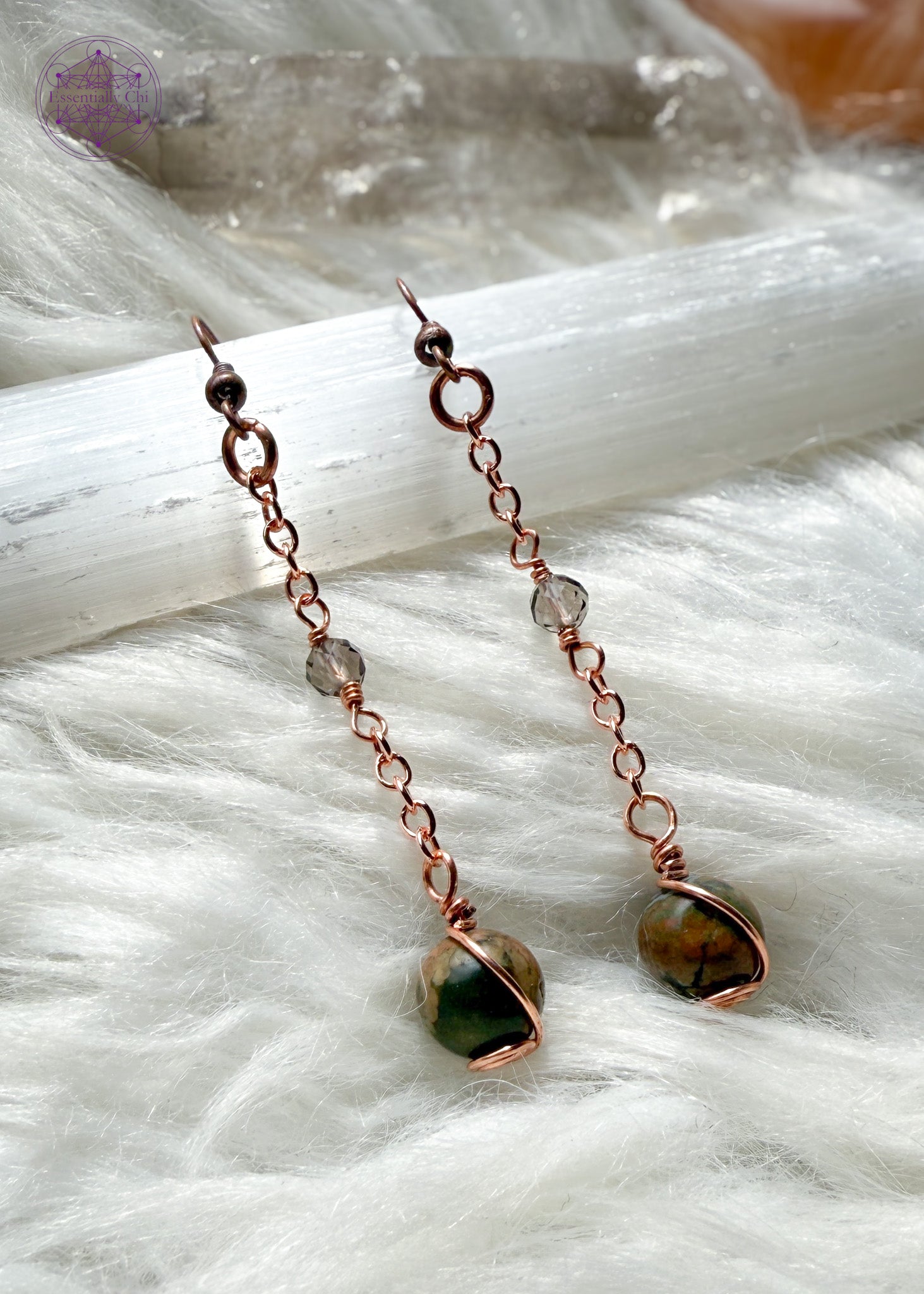 Gorgeous earthy rainforest rhyolite dangle earrings hang from a long copper chain and swirled in copper wire with smoky quartz faceted accent bead hung in the middle of the copper chain. Hung on brass ear hooks gives them a total length of 2.5". Shown on a white faux fur and selenite crystals around it.