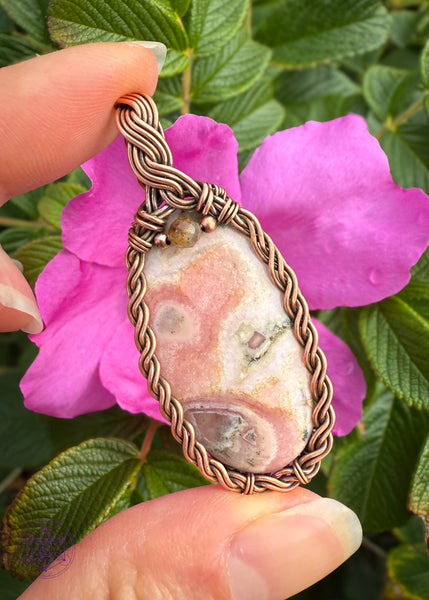 Copper wire wrapped pendant with an oval rhodochrosite crystal in a braided frame and a olive colored tourmaline crystal at the base of the bail. Held in hand in front of a pink wild rose to help the pinks in the crystal stand out. 