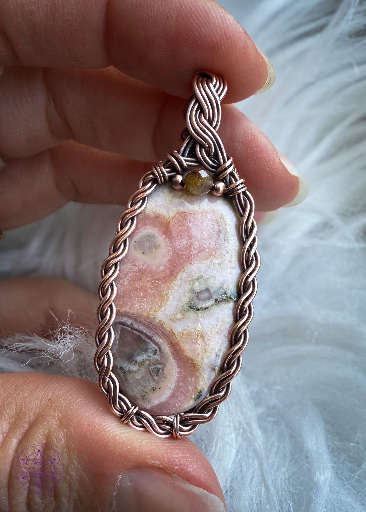 Copper wire wrapped pendant with an oval rhodochrosite crystal in a braided frame and a olive colored tourmaline crystal at the base of the bail. Held in hand in front of a faux white fur to show the details in the pendant.