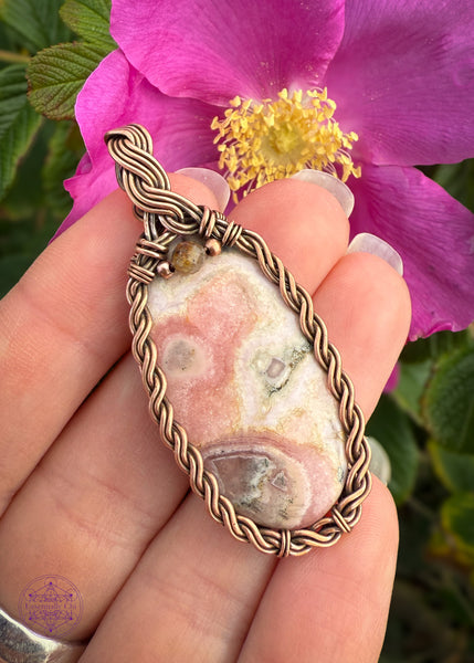 Copper wire wrapped pendant with an oval rhodochrosite crystal in a braided frame and a olive colored tourmaline crystal at the base of the bail. Held in hand  to show size reference in front of a pink wild rose to help the pinks in the crystal stand out. 