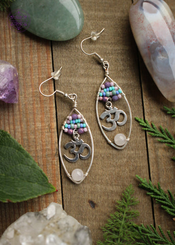 Rose quartz oval hoop earrings with a pastel beaded top of 4 lines of glass beads in colors of pink, purple, and turquoise and an om sign hanging from the 4th loine of beads. At the bottom of the hoop is a light pink rose quartz bead. These are so pretty wrapped on a silver plated wire hoop. Sitting on a wooden table with crystals and green foliage around it.