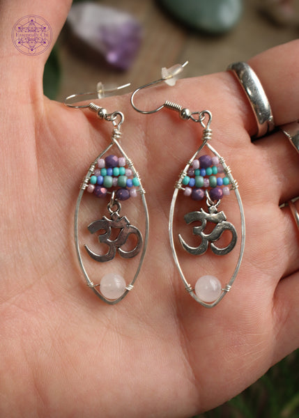 Rose quartz oval hoop earrings with a pastel beaded top of 4 lines of glass beads in colors of pink, purple, and turquoise and an om sign hanging from the 4th loine of beads. At the bottom of the hoop is a light pink rose quartz bead. These are so pretty wrapped on a silver plated wire hoop. Held in hand to show size reference.