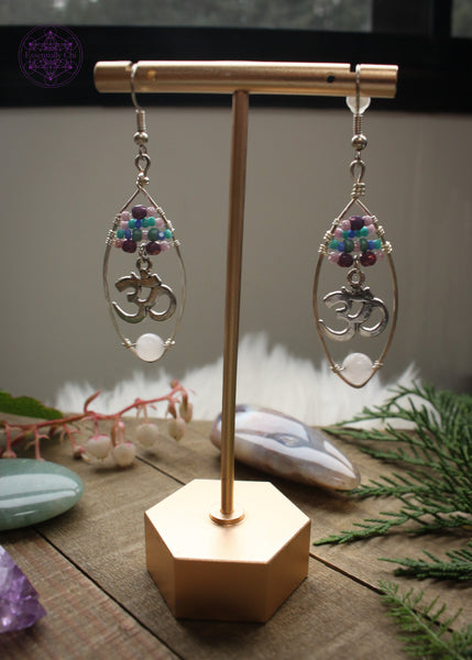 Rose quartz oval hoop earrings with a pastel beaded top of 4 lines of glass beads in colors of pink, purple, and turquoise and an om sign hanging from the 4th loine of beads. At the bottom of the hoop is a light pink rose quartz bead. These are so pretty wrapped on a silver plated wire hoop. Shown hanging from earring stand and the light coming through behind them. 