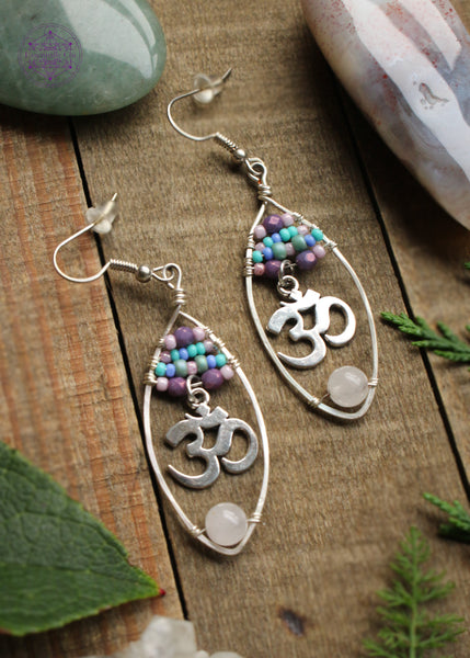 Rose quartz oval hoop earrings with a pastel beaded top of 4 lines of glass beads in colors of pink, purple, and turquoise and an om sign hanging from the 4th loine of beads. At the bottom of the hoop is a light pink rose quartz bead. These are so pretty wrapped on a silver plated wire hoop. Sitting on a wooden table with crystals and green foliage around it.