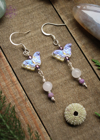 These dangle earrings have an iridescent acrylic butterfly followed by beads of rose quartz, phosphosiderite, and glass beads in frosty white and purple.  Hung on sterling silver ear hooks these earrings are so fairylike and whimsy! Sitting on a wooden table with crystals and foliage around it.