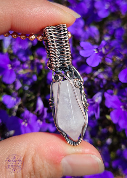 handcrafted filagree crystal necklace with a small rose quartz point wrapped in copper wire. Held in hand over purple flowers. Showing the back which is just as decorative as the front.