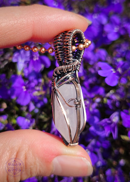 handcrafted filagree crystal necklace with a small rose quartz point wrapped in copper wire. Held in hand over purple flowers. Showing right side.