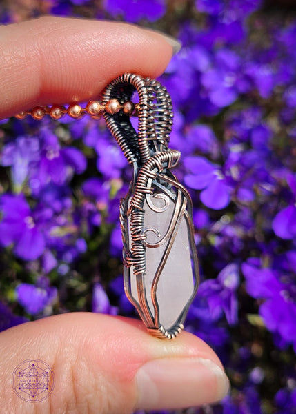 handcrafted filagree crystal necklace with a small rose quartz point wrapped in copper wire. Held in hand over purple flowers. Showing the left.