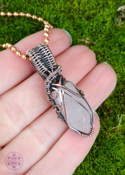 handcrafted filagree crystal necklace with a small rose quartz point wrapped in copper wire. Held in hand for size reference over green moss.