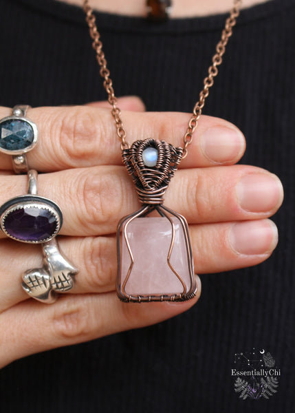 Love Potion #9 Pendant - A copper wire-wrapped pendant featuring Rose Quartz and Rainbow Moonstone. Symbolizing love, emotional healing, and intuition. Ideal for Gemini, Cancer, and Libra. Handcrafted with an enchanting design. The rose quartz is a tapered square cut and the copper wire is simple to hug the stone, the bale is a channel setting with a flashy blue small round rainbow moonstone. Shown worn and how it hangs along the neckline.