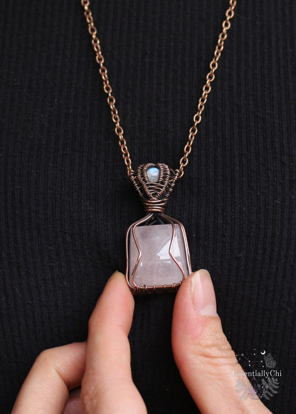 Love Potion #9 Pendant - A copper wire-wrapped pendant featuring Rose Quartz and Rainbow Moonstone. Symbolizing love, emotional healing, and intuition. Ideal for Gemini, Cancer, and Libra. Handcrafted with an enchanting design. The rose quartz is a tapered square cut and the copper wire is simple to hug the stone, the bale is a channel setting with a flashy blue small round rainbow moonstone. Shown on the body holding it with my fingers for size reference. 