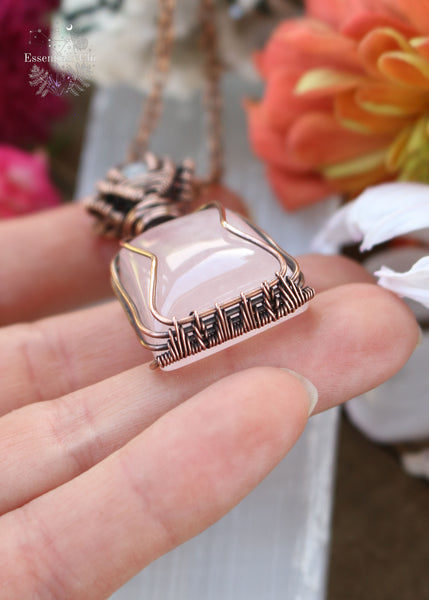 Love Potion #9 Pendant - A copper wire-wrapped pendant featuring Rose Quartz and Rainbow Moonstone. Symbolizing love, emotional healing, and intuition. Ideal for Gemini, Cancer, and Libra. Handcrafted with an enchanting design. The rose quartz is a tapered square cut and the copper wire is simple to hug the stone, the bale is a channel setting with a flashy blue small round rainbow moonstone. Showing the bottom weaving pattern. 