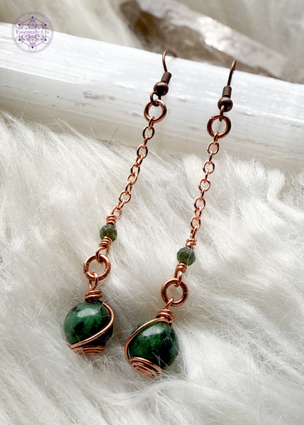 Gorgeous deep green Ruby Zoisite dangle earrings hang from a long copper chain and swirled in copper wire with green tourmaline faceted accent bead. Hung on brass ear hooks gives them a total length of 2.75". Shown on selenite and white faux fur and crystals.