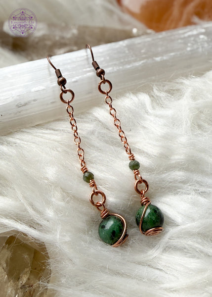 Gorgeous deep green Ruby Zoisite dangle earrings hang from a long copper chain and swirled in copper wire with green tourmaline faceted accent bead. Hung on brass ear hooks gives them a total length of 2.75". Shown on selenite and white faux fur and crystals.