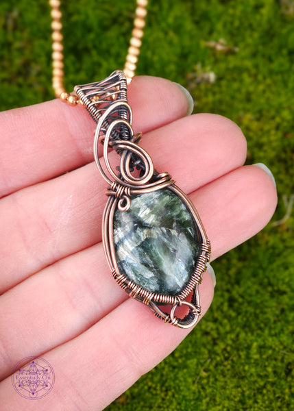 feathery green oval seraphinite crystal wrapped in copper wire for an elegant everyday necklace. There is a small loop at the bottom to hug around the bottom of the crystal and a few swirls along the bail. Hung on a copper ball and chain necklace. Held in hand for size refernce.