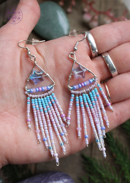 Shooting star fringe earrings. The design is a silver plated triangle with an blue/purple acrylic star in the middle and a fringe with a gradient of light blue pink and white hanging from the bottom of the triangle. Held in hand to show the size reference. (4" long including the stainless steel ear hooks).