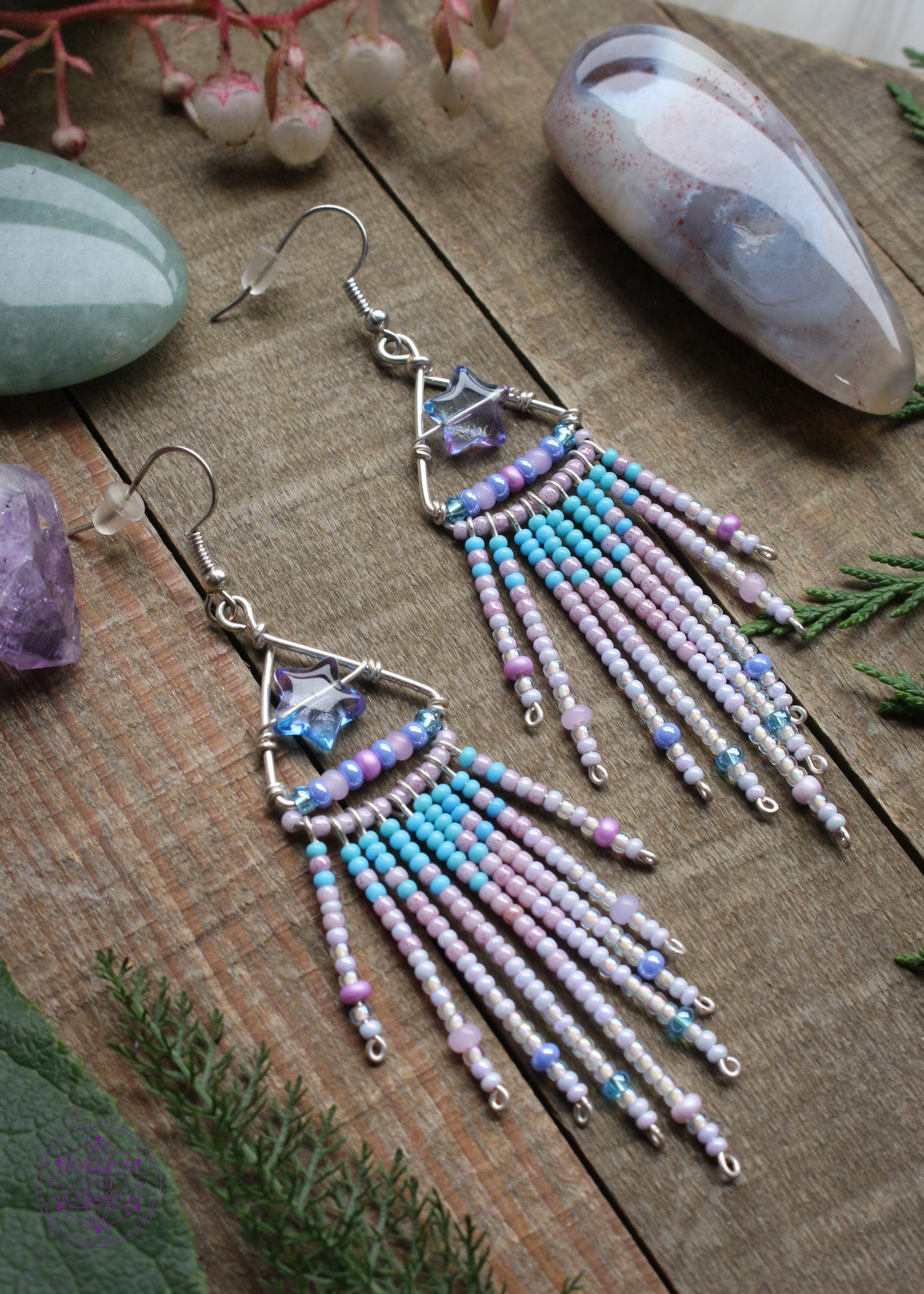 Shooting star fringe earrings. The design is a silver plated triangle with an blue/purple acrylic star in the middle and a fringe with a gradient of light blue pink and white hanging from the bottom of the triangle. Sitting on a wooden table with crystals and foliage around it.