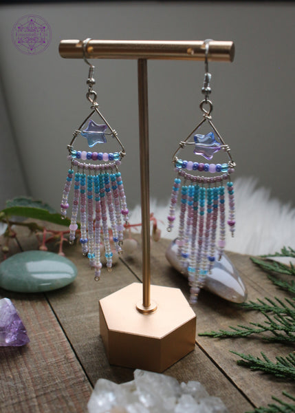 Shooting star fringe earrings hanging from an earring stand with the light behind it shining through. The design is a silver plated triangle with an blue/purple acrylic star in the middle and a fringe with a gradient of light blue pink and white hanging from the bottom of the triangle. Sitting on a wooden table with crystals and foliage around it.