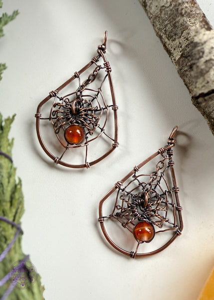copper wire wrapped spider web earrings shown laying flat on the table. The web is in a tear drop shape and the spider web dangles from the top over the middle of the web. The spider can have his legs attach to the web if you prefer it not to dangle. Hangs on sterling silver ear hooks.