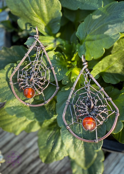 copper wire wrapped spider web earrings shown laying on green leaves in natural light. The web is in a tear drop shape and the spider web dangles from the top over the middle of the web. The spider can have his legs attach to the web if you prefer it not to dangle. Hangs on sterling silver ear hooks.