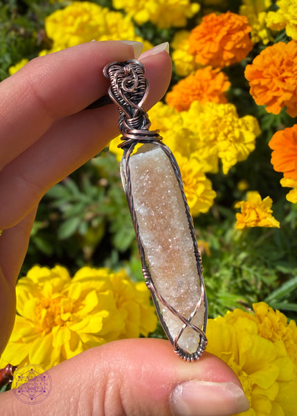 Spirit Quartz point crystal pendant wrapped in copper wire. A small criss cross along the bottom of the point to hold the stone secure with twisted square wire along the sides to catch the light. The bail has a swirl along the front and texture along the top. Shown in hand with yellow and orange marigolds in the background.