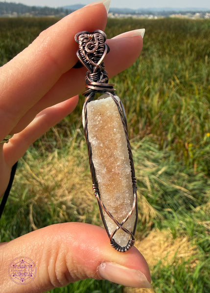 Spirit Quartz point crystal pendant wrapped in copper wire. A small criss cross along the bottom of the point to hold the stone secure with twisted square wire along the sides to catch the light. The bail has a swirl along the front and texture along the top. Shown in hand grassy wetlands in the background.