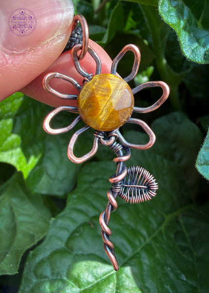 Copper wire wrapped sunflower pendant with a yellow tiger eye faceted bead in the center of the flower. A wire woven leaf is attached three-quarters up the stem. Held in hand with dark green leaves from my garden behind it.