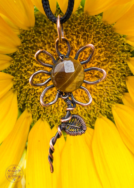Copper wire wrapped sunflower pendant with a yellow tiger eye faceted bead in the center of the flower. A wire woven leaf is attached three-quarters up the stem. Hanging in front of a yellow sunflower. 