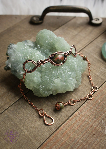 Copper wire wrapped bracelet features an oval Unakite bead wrapped in twisted copper design with loops on each end. Shown on a green crystal to show all the details in the center, sides and clasp. 