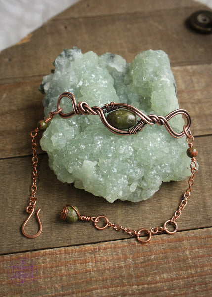 Copper wire wrapped bracelet features an oval Unakite bead wrapped in twisted copper design with loops on each end. Shown on a green crystal to show all the details in the center, sides and clasp. The middle bead rotates and on one side it's green and pink on the other is a mossy green.