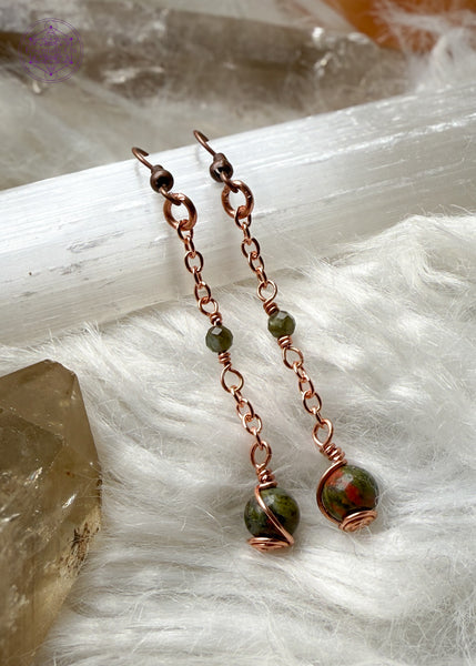 Gorgeous earthy green Unakite dangle earrings hang from a long copper chain and swirled in copper wire with green tourmaline faceted accent bead hung in the middle of the copper chain. Hung on brass ear hooks gives them a total length of 2.5". Shown on white faux fur with crystals around it.