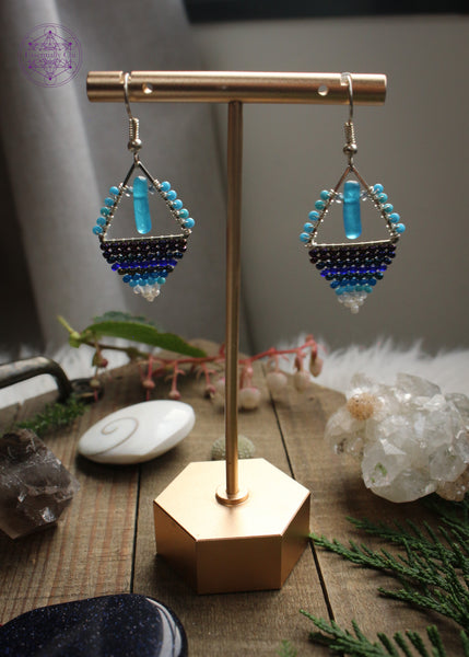 Beaded earrings in a diamond shape features a glass cylinder danging from the top middle and a gradient of dark blue light blue to white. Hung on stainless steel ear hooks. Hanging on an earring stand to let the light behind it shine through on a wooden table with crystals and foliage around it.