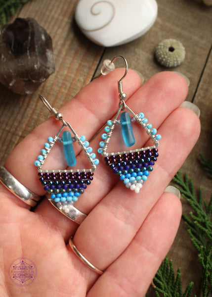 Beaded earrings in a diamond shape features a glass cylinder danging from the top middle and a gradient of dark blue light blue to white. Hung on stainless steel ear hooks. Held in hand to show size reference of 2".