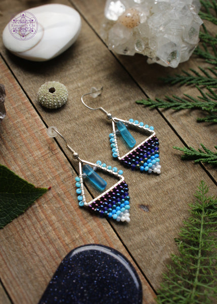 Beaded earrings in a diamond shape features a glass cylinder danging from the top middle and a gradient of dark blue light blue to white. Hung on stainless steel ear hooks. Sitting on a wooden table with crystals and foliage around it.