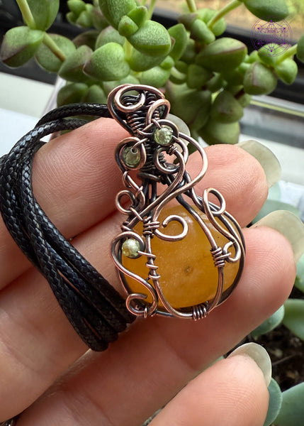 Orange Aventurine mini pumpkin pendant with swirls coming down from the bail and over the front of the pumpkin with three peridot beads in the swirls to act as leaves. Shown indoors for different lighting. 
