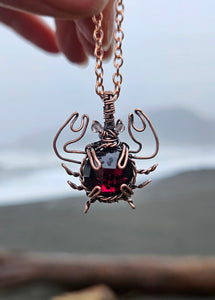 Copper wire wrapped crab necklace with a round faceted garnet crystal and small faceted smoky quartz beads for eyes. The legs are twisted to make them thin while the pincher arms are bigger to represent actual crab anatomy. Held up in front of the ocean waves on a stormy day.