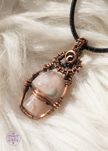 Copper wire wrapped flower agate mushroom pendant with copper swirls along the top and a small cage around the mushroom to keep it stable. The mushroom moves freely like a fidget spinner. Shown resting on a white faux fur background.