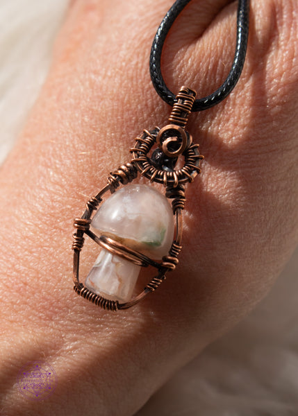 Copper wire wrapped flower agate mushroom pendant with copper swirls along the top and a small cage around the mushroom to keep it stable. The mushroom moves freely like a fidget spinner. Shown on the back of my hand.