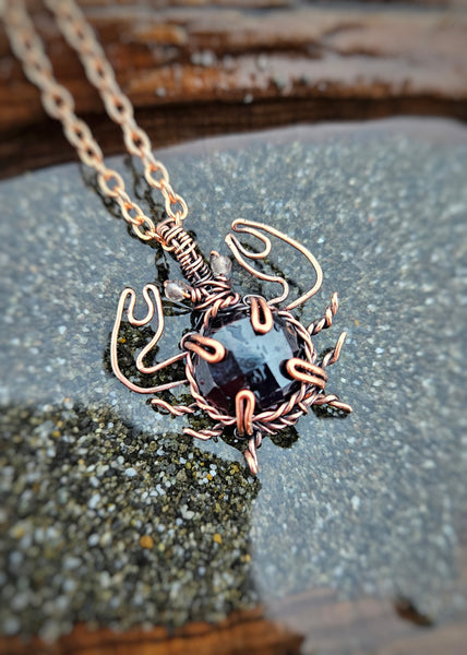 Copper wire wrapped crab necklace with a round faceted garnet crystal and small faceted smoky quartz beads for eyes. The legs are twisted to make them thin while the pincher arms are bigger to represent actual crab anatomy. Laying in a small tide pool with sand to simulate a sunken treasure found on the beach.