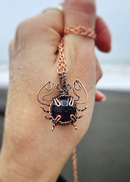 Copper wire wrapped crab necklace with a round faceted garnet crystal and small faceted smoky quartz beads for eyes. The legs are twisted to make them thin while the pincher arms are bigger to represent actual crab anatomy. Held up against my hand for size refrence.