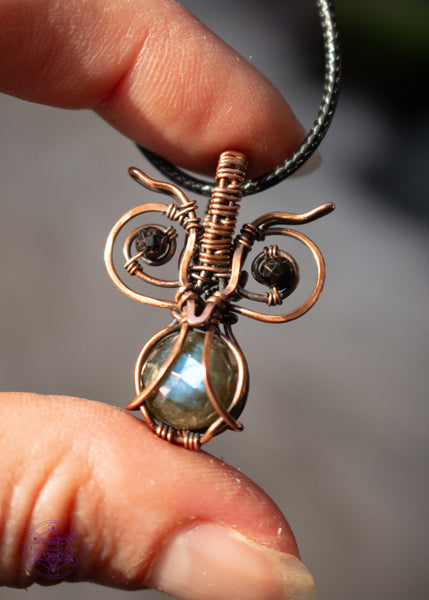 copper wire wrapped pygmy owl pendant with a faceted blue flash labradorite belly and dark green tourmaline beads for eyes, shown held in hand with the pendant in the light.