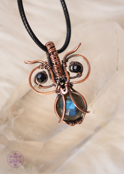 copper wire wrapped pygmy owl pendant with a faceted blue flash labradorite belly and dark green tourmaline beads for eyes, shown on a clear quartz crystal and a white background.