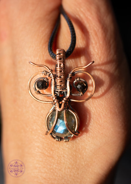 xcopper wire wrapped pygmy owl pendant with a faceted blue flash labradorite belly and dark green tourmaline beads for eyes, shown held on the back of my hand with a contrast of light and dark.